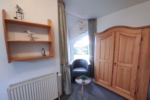 A room with a radiator, a chair, a small table with a plant, a wooden wardrobe, and a shelf with decorative items. - Oysterfinders (1230) (Neßmersiel)