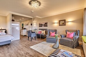 Ein Hotelzimmer mit einem Bett, zwei gestreiften Sesseln, einem Esstisch mit Stühlen, einer Kristallleuchtre und Kunstwerken an den Wänden.