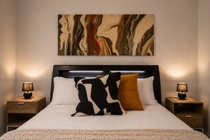A neatly made bed with a headboard, two bedside tables with lamps, and a large abstract painting above the bed. - Docklands Rooftop Pool Retreat (Docklands)