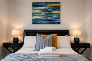 A neatly made bed with white and striped pillows, a rolled white towel, and a blue blanket. There are two bedside tables with lamps and a painting above the bed. - Docklands Rooftop Pool Retreat (Docklands)