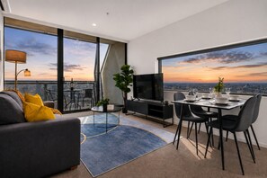 A modern living room with a sofa, TV, dining table, and chairs, large windows with a city view, and a balcony with a table and chairs. - Flagstaff Park View 2 Bedroom (Melbourne)