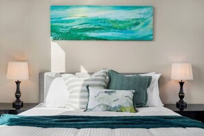 A neatly made bed with striped bedding, a teal throw blanket, and a painting of a blue ocean above it. - Flagstaff Park View 2 Bedroom (Melbourne)