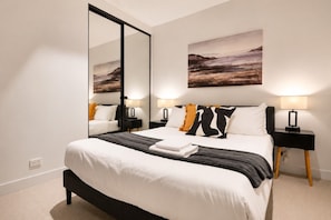 A modern hotel room with a large bed, two bedside tables with lamps, a large mirror, and a landscape painting on the wall. - Blue Haven Docklands 2 Bedroom (Docklands)