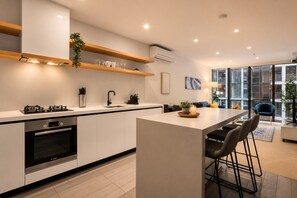 A modern kitchen with a central island, built-in oven, and a dining area with bar stools. - Blue Haven Docklands 2 Bedroom (Docklands)
