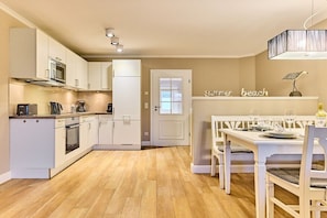 A modern kitchen with white cabinetry, a dining area with a white table and chairs, and a 'summer beach' sign on the wall. - Holiday apartment with 2 bedrooms (Westerland)