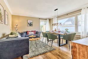 A modern living room with a sofa, dining table, chairs, and a coffee table. There is a window with curtains, a floor lamp, and artwork on the walls. - Holiday apartment with 3 bedrooms (Wenningstedt)