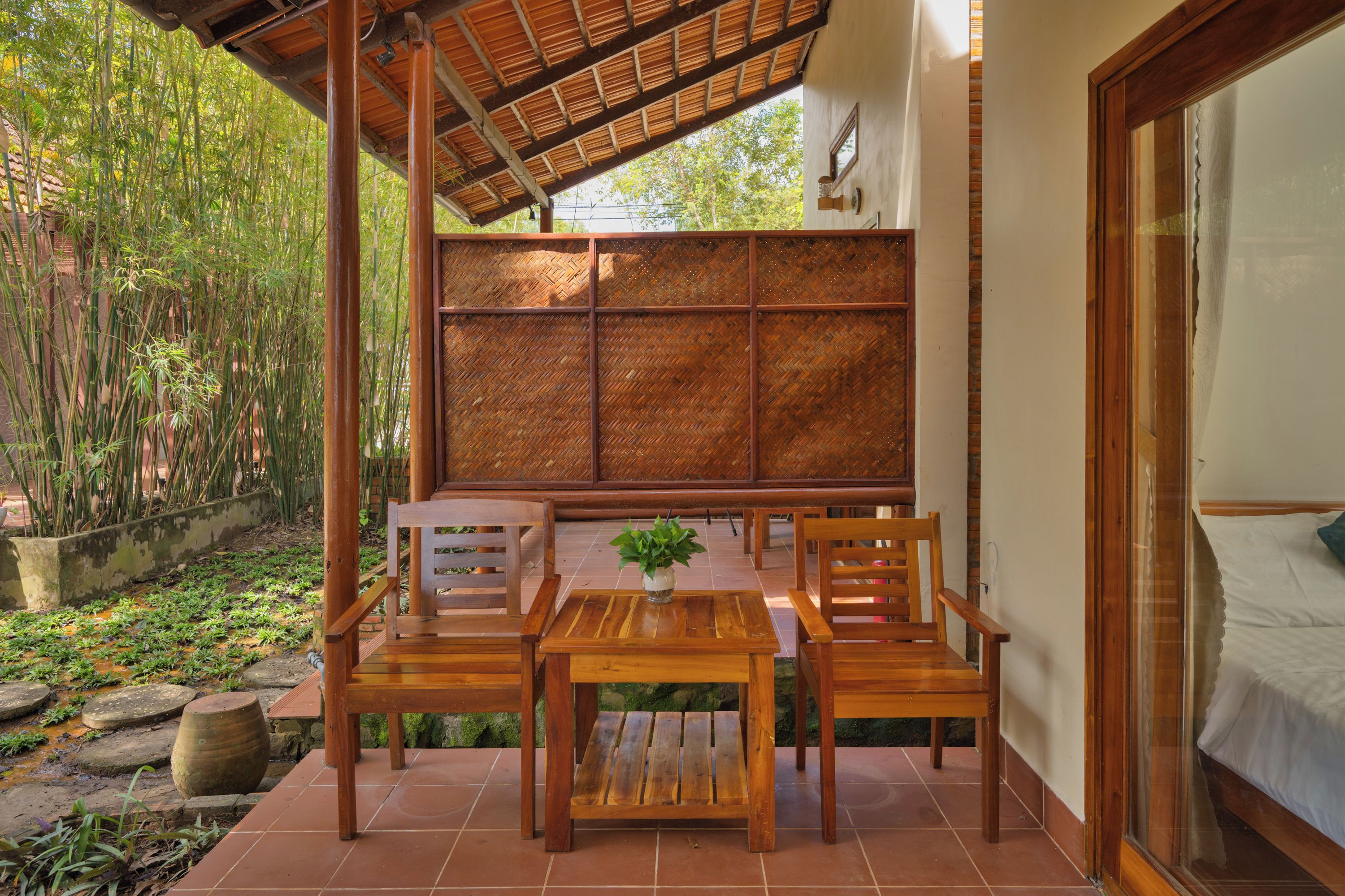 Un área de estar al aire libre con muebles de madera, una mesa, sillas y un fondo de bambú, con vista a un bosque de bambú.
