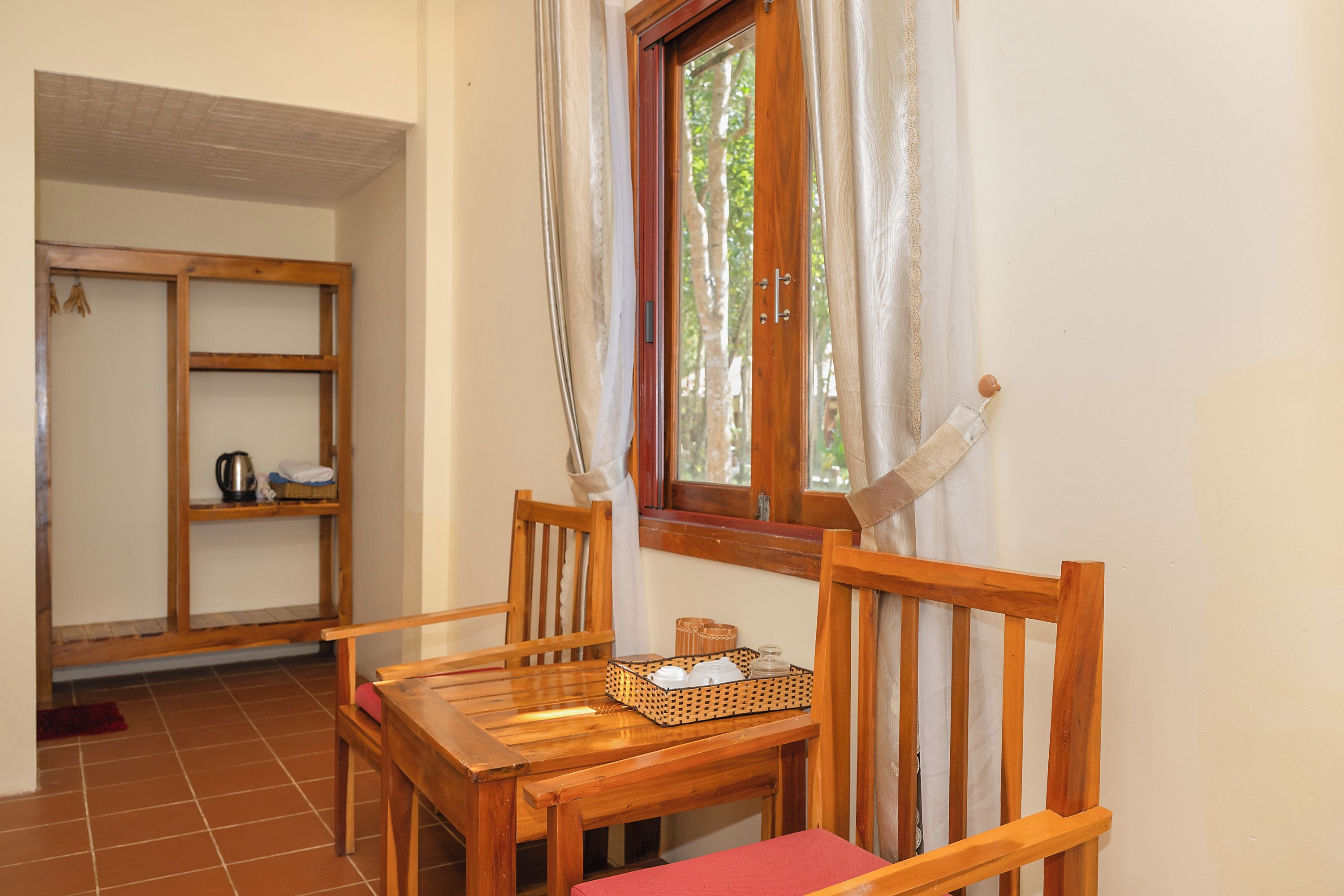 A wooden dining table with chairs, a window with curtains, and a shelf with kitchen items.