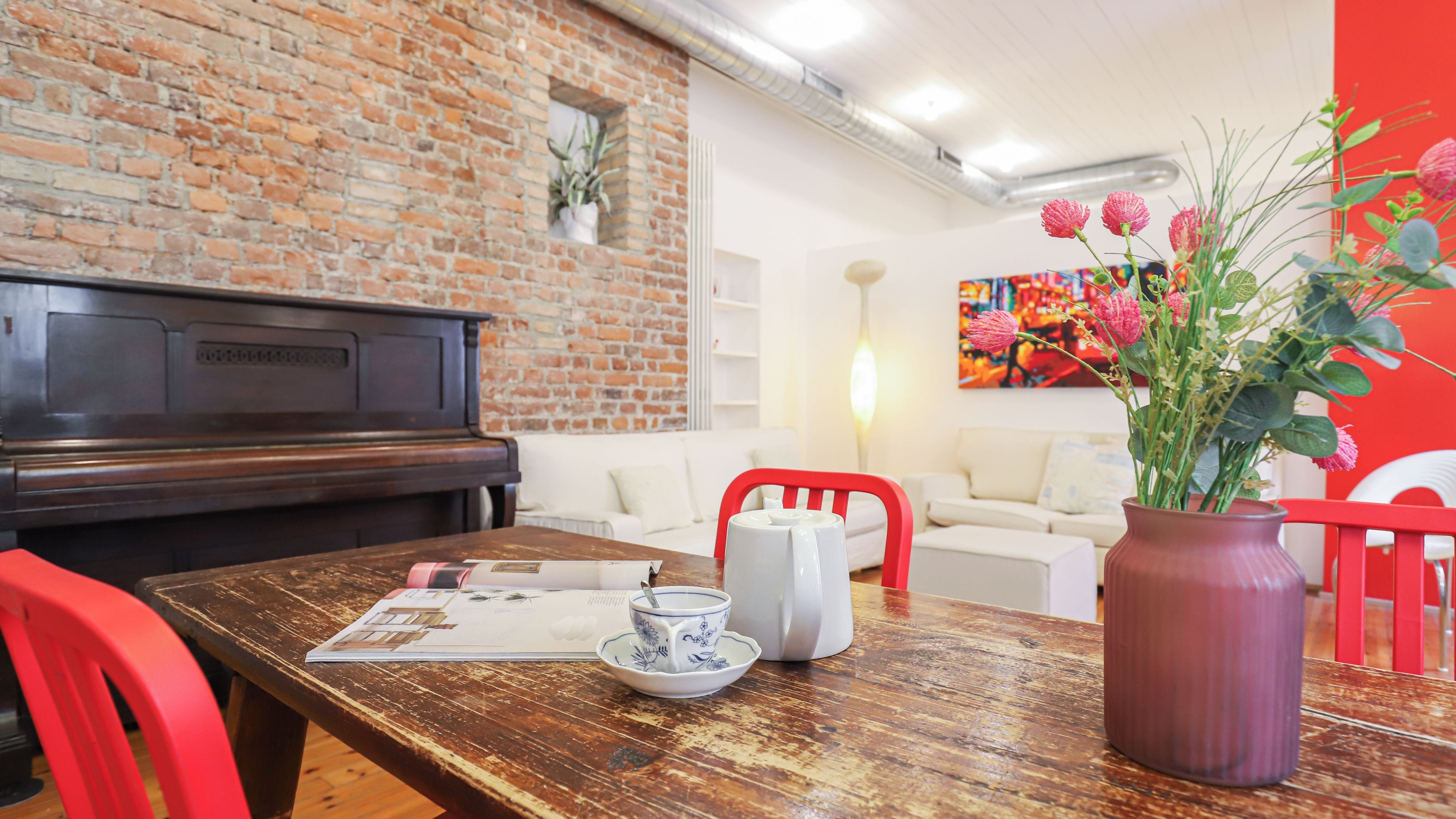 A room with a brick wall, a piano, a wooden table with a cup and saucer, a red chair, and a vase with flowers.