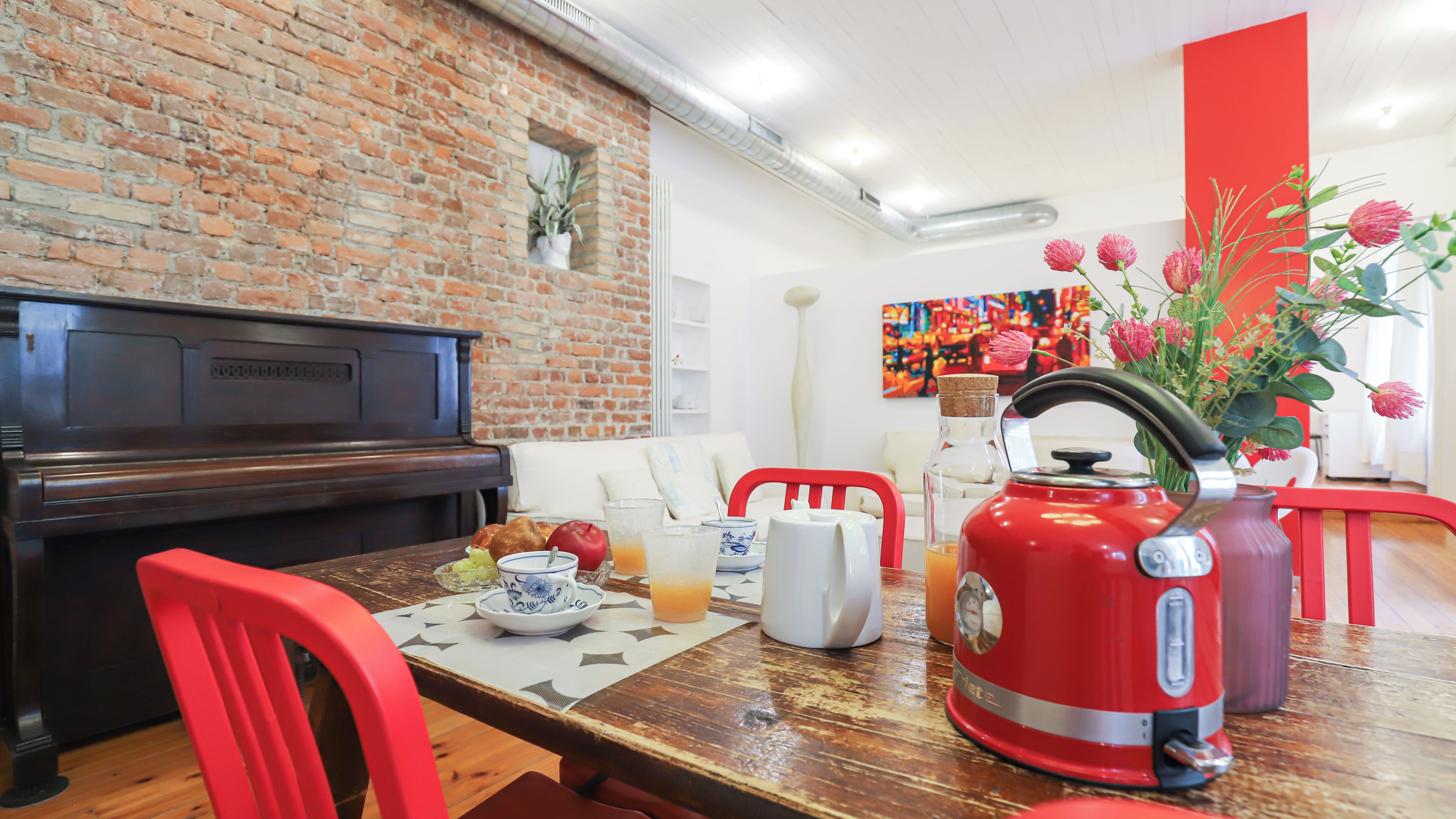 Un comedor con pared de ladrillo, un piano, un hervidor rojo, y una mesa servida con platos, vasos y un jarrón de flores.