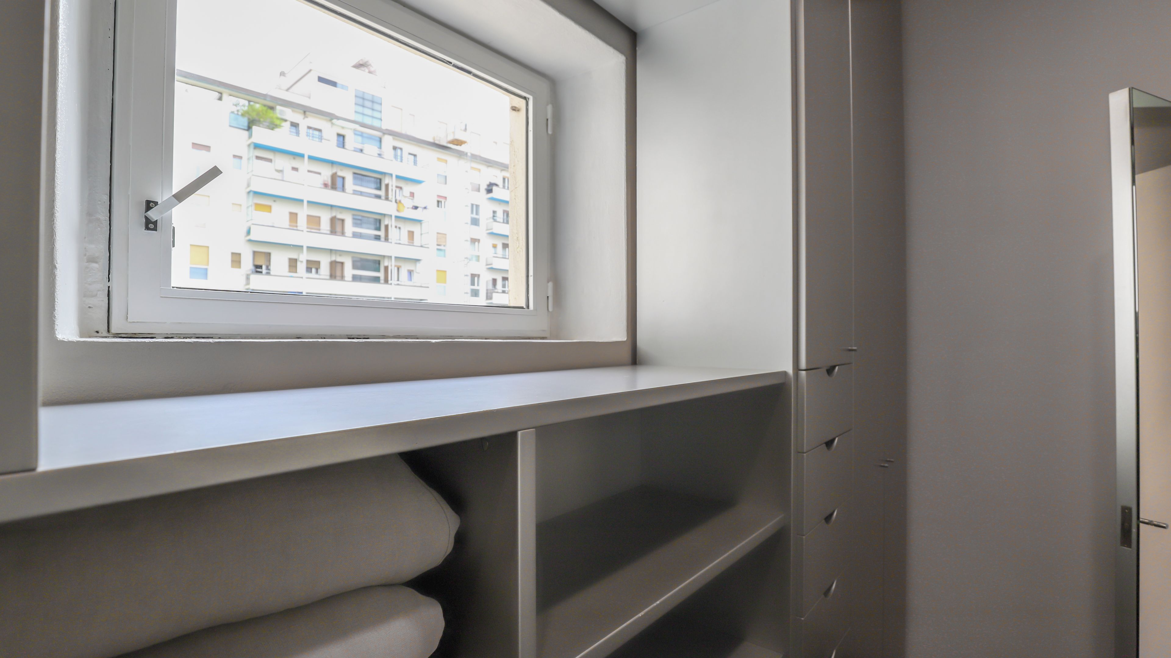 Una habitación con ventana que da a un edificio de varios pisos, estanterías empotradas y una puerta.