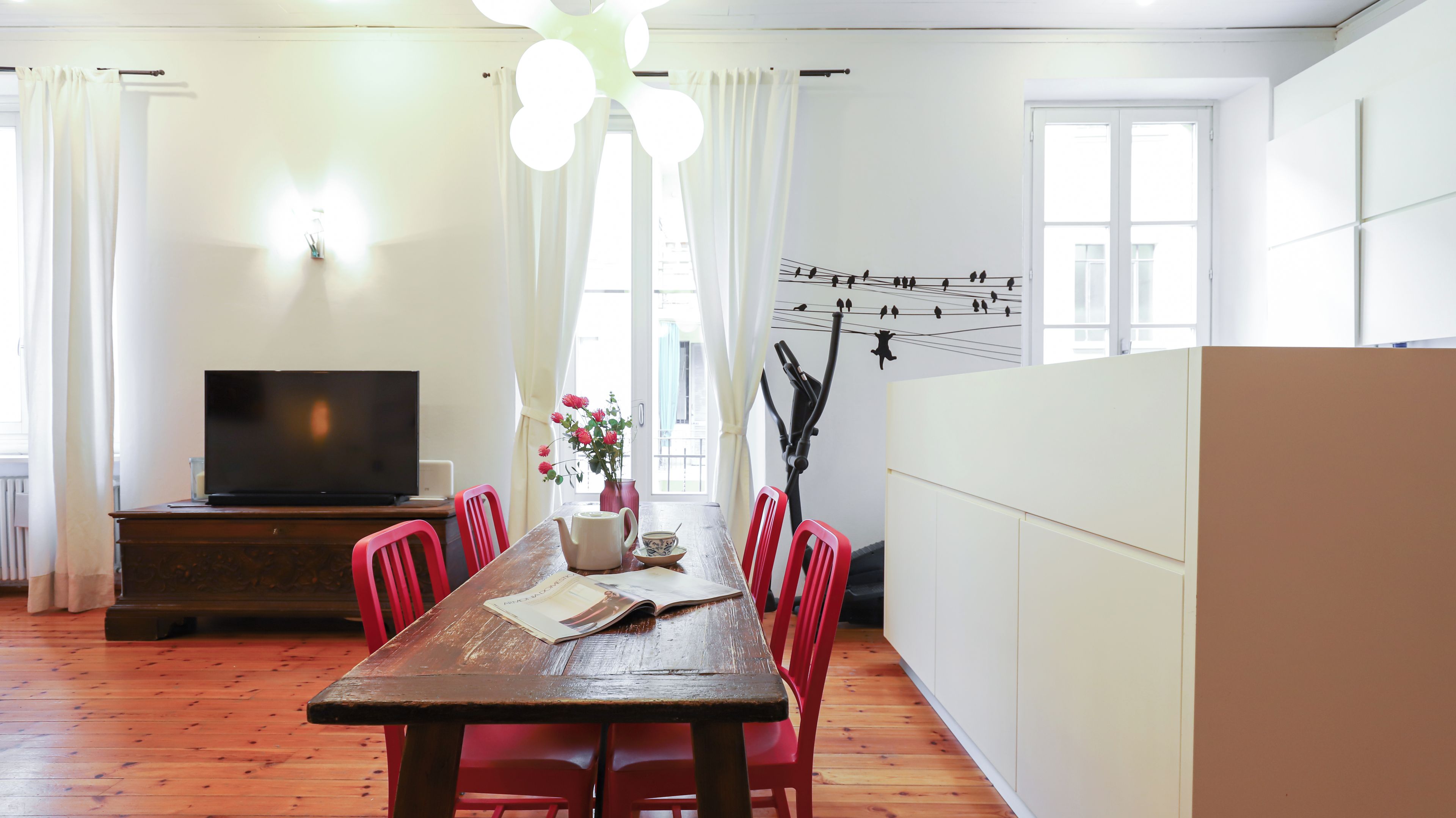Un comedor con una mesa de madera, sillas rojas, un televisor y una pared decorada con un mural de pájaros.