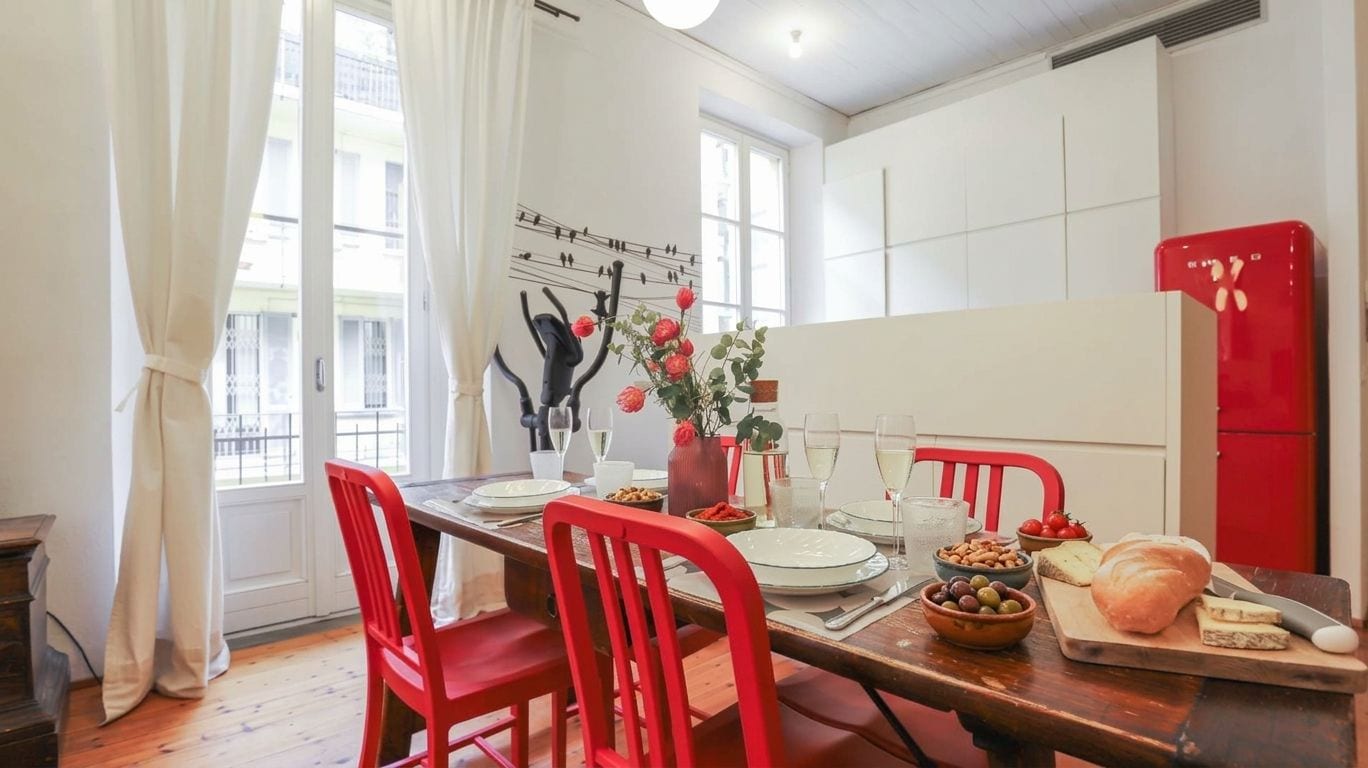 A dining area with a wooden table set for four, red chairs, a red refrigerator, and a window with curtains.