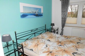 A bedroom with a bed, a nightstand, a lamp, a window with curtains, and a painting on the wall. - Holiday apartment with 1 bedroom (18225 Kühlungsborn, Deutschland)