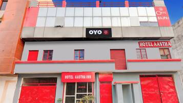 Ett hotell med en röd och grå fasad, en tydlig skylt med texten ”Hotel Aastha Inn” och en väl synlig ”OYO”-logotyp.