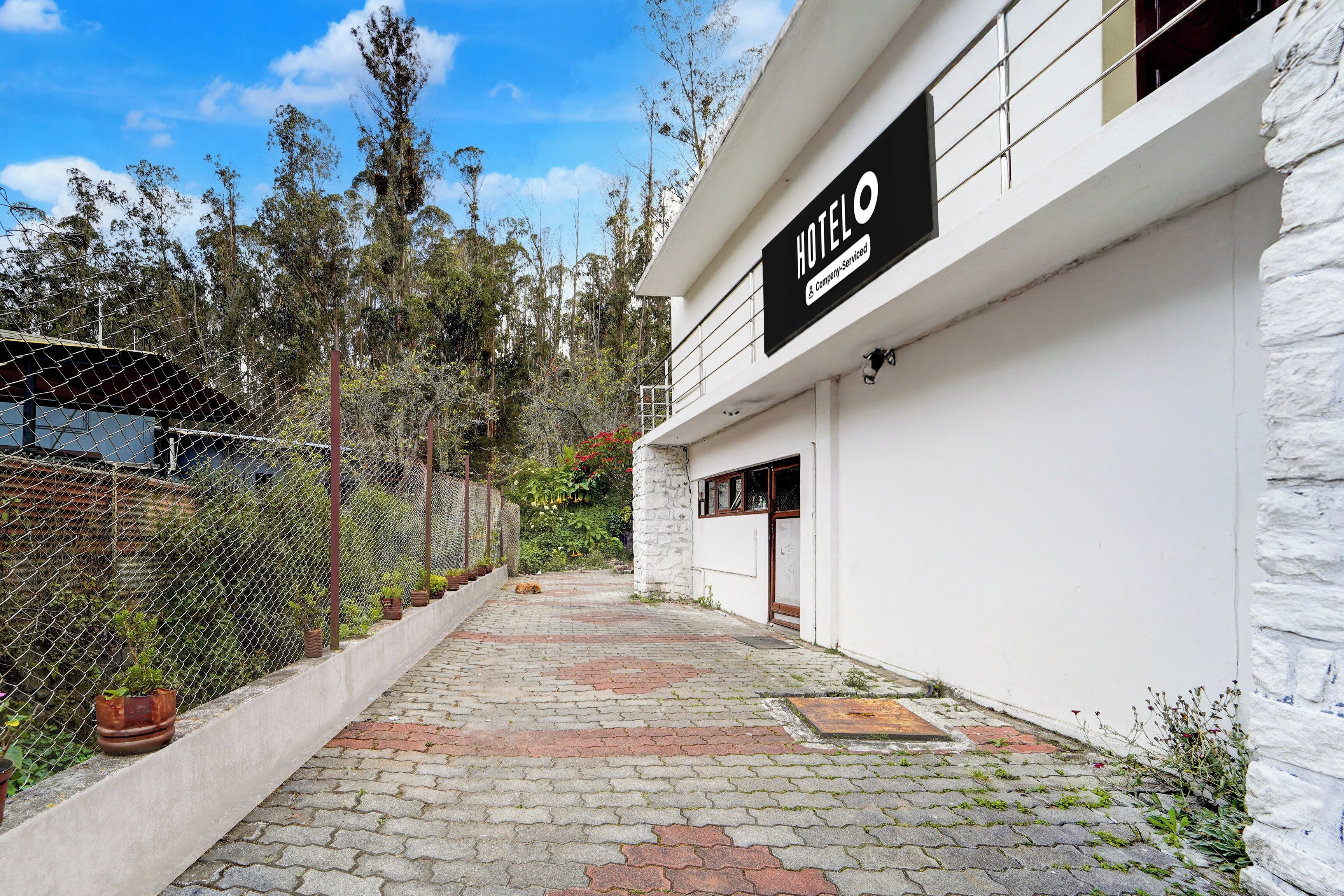 A hotel entrance with a paved pathway leading to the door, surrounded by a chain-link fence and vegetation.