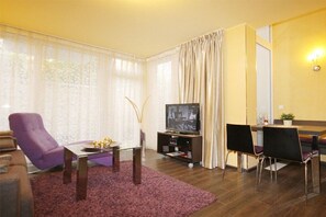 Olohuoneessa on sohva, violetti nojatuoli, sohvapöytä, jalustalla oleva televisio, ruokailutila, jossa on pöytä ja tuolit, sekä keittiötila, jossa on työtaso ja kasvi.