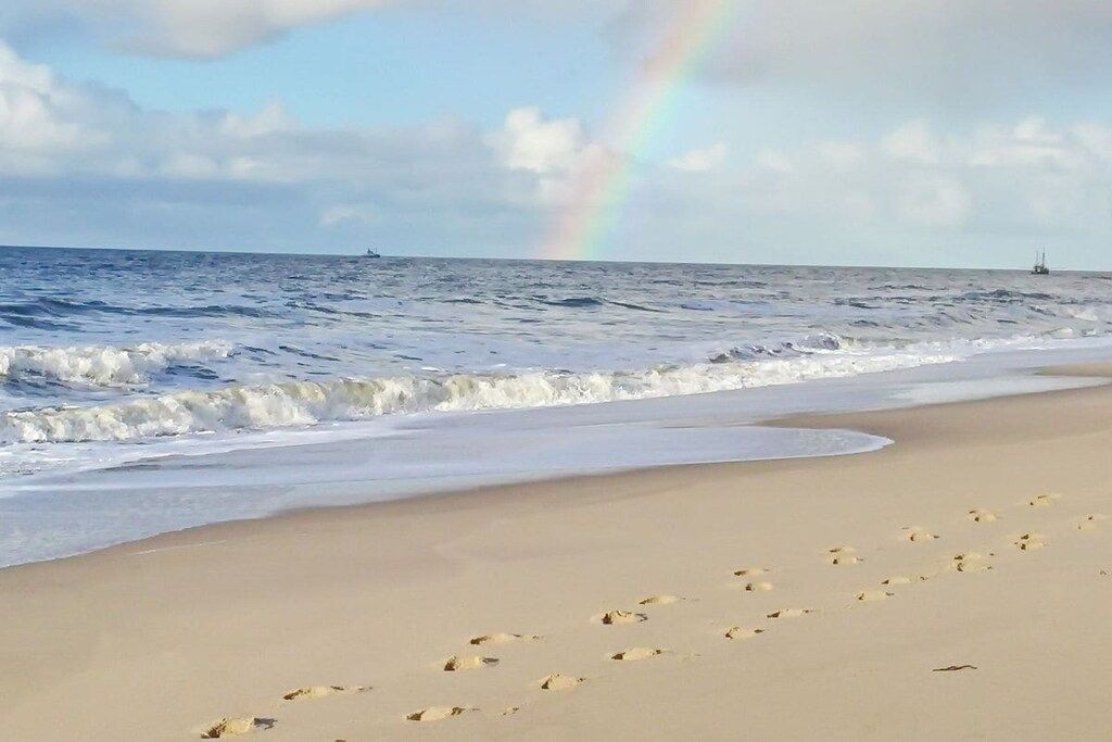 A beach with footprints leading to the water, a rainbow in the sky, and a ship in the distance.