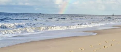 A beach with footprints leading to the water, a rainbow in the sky, and a ship in the distance.