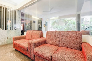 Uma área de recepção com divisória de vidro, balcão de recepção em mármore e sofás vermelhos com padrão.