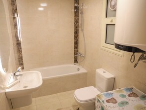 A bathroom with a bathtub, sink, toilet, and a window with a mirror. - Your Dream Studio in New Cairo E104, AC, Wi-Fi, Prime Location (Cairo Governorate)