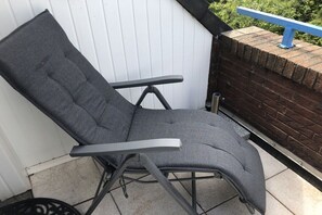Ein grauer Außenstuhl mit Metallgestell, platziert auf einer gefliesten Terrasse neben einer weißen Wand und einer Ziegelwand mit blauer Brüstung.