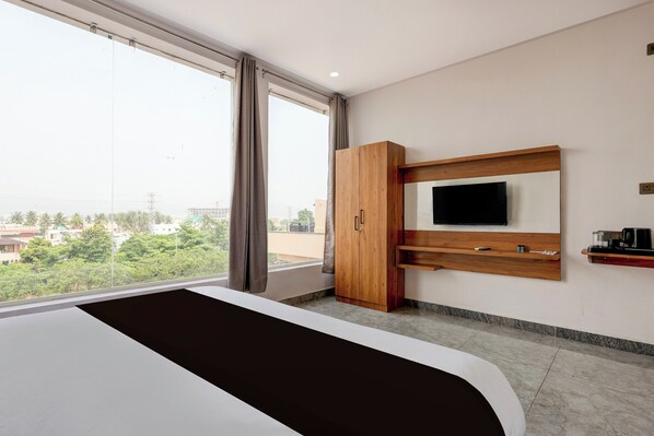 A hotel room with a large bed, a TV mounted on a wooden shelf, and a view of the cityscape through the window.