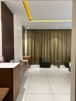 A modern hotel room with a reception desk, a green sofa, a glass table, and a large window with curtains. - MEHRAN (Vijayapura)