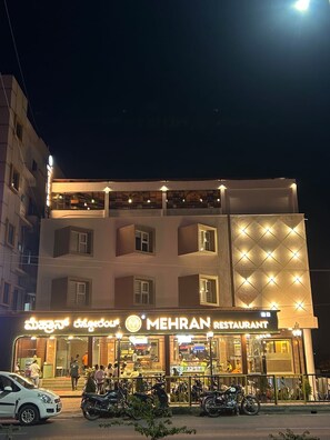 A multi-story building with a restaurant named 'MEHRAN RESTAURANT' on the ground floor. There are several motorcycles parked in front, and a white car is visible. - MEHRAN (Vijayapura)