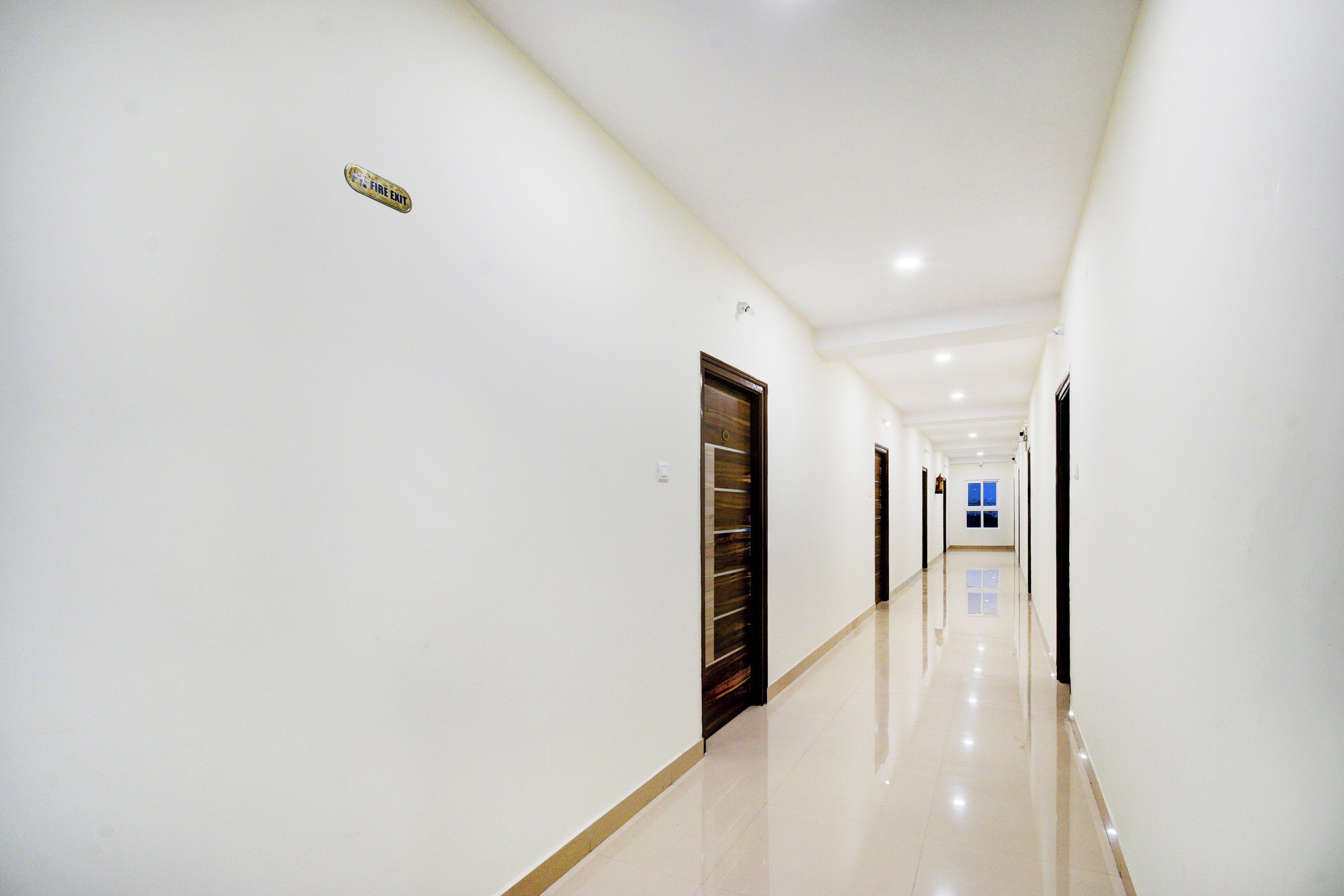 A long, well-lit hallway with doors on one side and a fire exit sign on the wall.