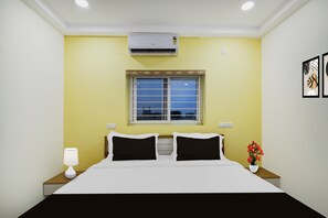 A hotel room with a bed, a window, an air conditioner, a nightstand with a lamp, and a wall-mounted phone.