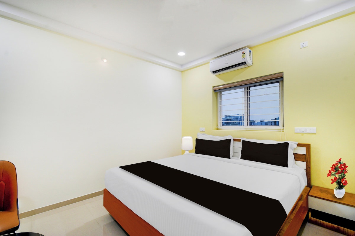 A hotel room with a bed, a nightstand, a lamp, an air conditioning unit, and a window with a view.
