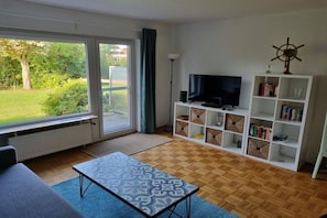 A living room with a television, bookshelf, and a view of the outdoors through a sliding glass door. - Holiday apartment with 1 bedroom (Norddeich)