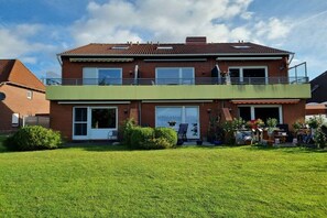 A two-story residential building with a flat roof, a balcony on the upper floor, and a front door with a small porch area. - Holiday apartment with 1 bedroom (Norddeich)