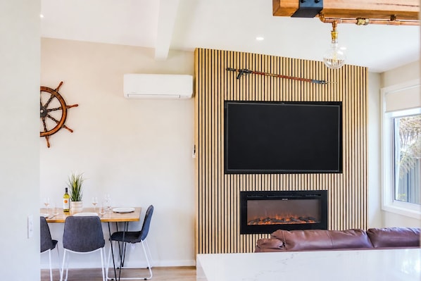 A modern living room with a fireplace, a mounted television, and a dining area with chairs and a table. - High-End Luxury Unit 8min Walk to Town & Cafes (Mallacoota)