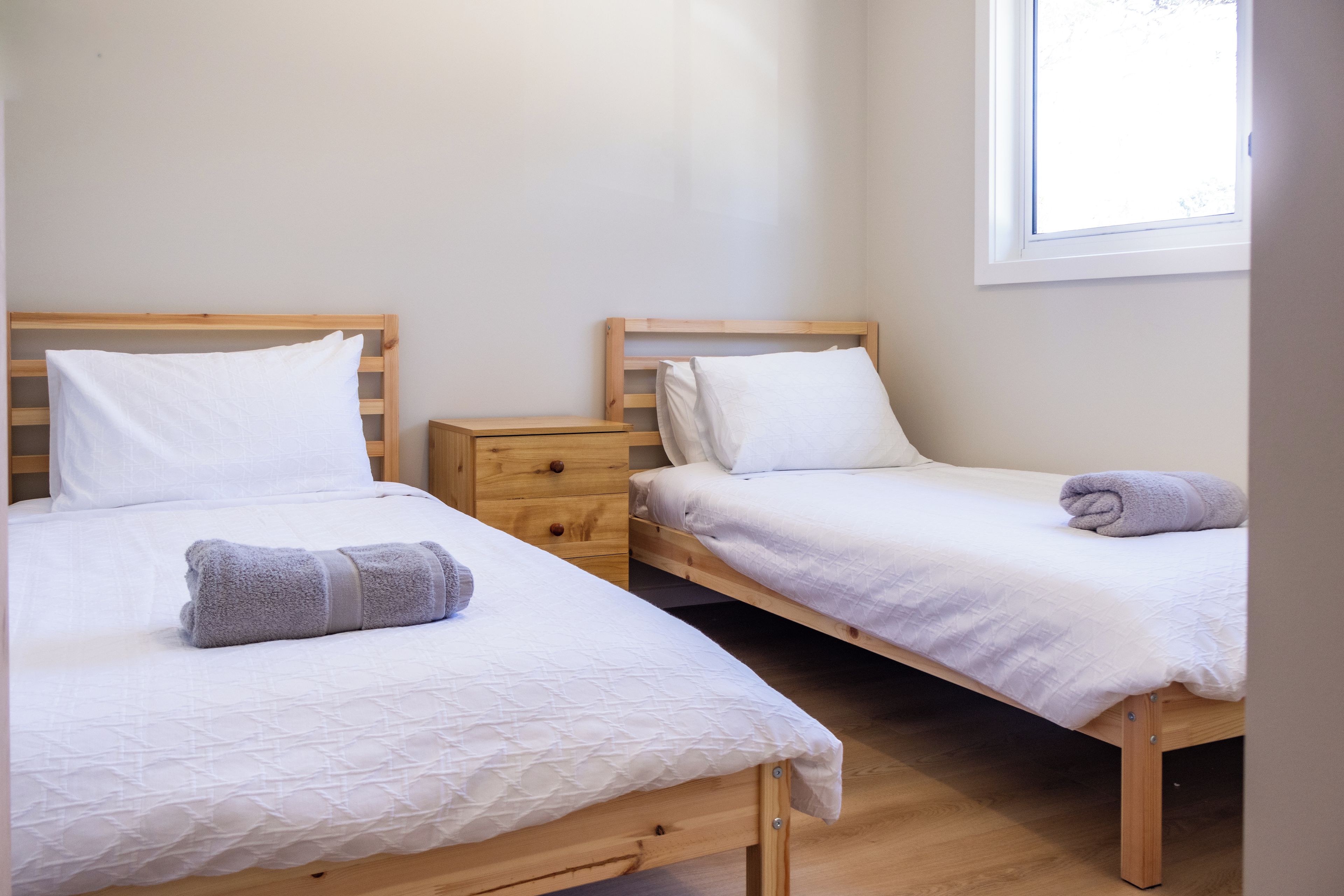 A room with two wooden beds, each with white bedding and pillows, a wooden nightstand, and a window providing natural light.