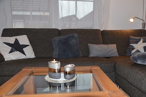 A modern living room with a dark grey sofa, two cushions with star patterns, a glass coffee table with candles, and a floor lamp. - Schürholz holiday apartment (Hooksiel)