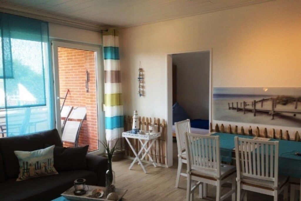 Une pièce comprenant un canapé, une petite table avec un vase et une chaise. Il y a une fenêtre avec des rideaux, une porte et un mur orné d’un tableau.
