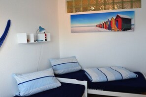 A bedroom with two beds, a painting of beach huts, a shelf with items, and a blue rope. - Holiday apartment Strandidyll (Horumersiel)