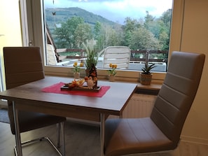 A dining area with a table and chairs, a window with a view of trees and hills, and potted plants on the windowsill. - Holiday Apartment 'Olsberg Panorama' with Mountain View and Private Balcony (Olsberg)