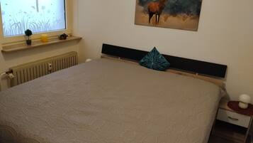 Un dormitorio con cama, ventana con vistas, radiador, mesita de noche y un cuadro en la pared.