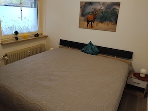 Un dormitorio con cama, ventana con vistas, radiador, mesita de noche y un cuadro en la pared.
