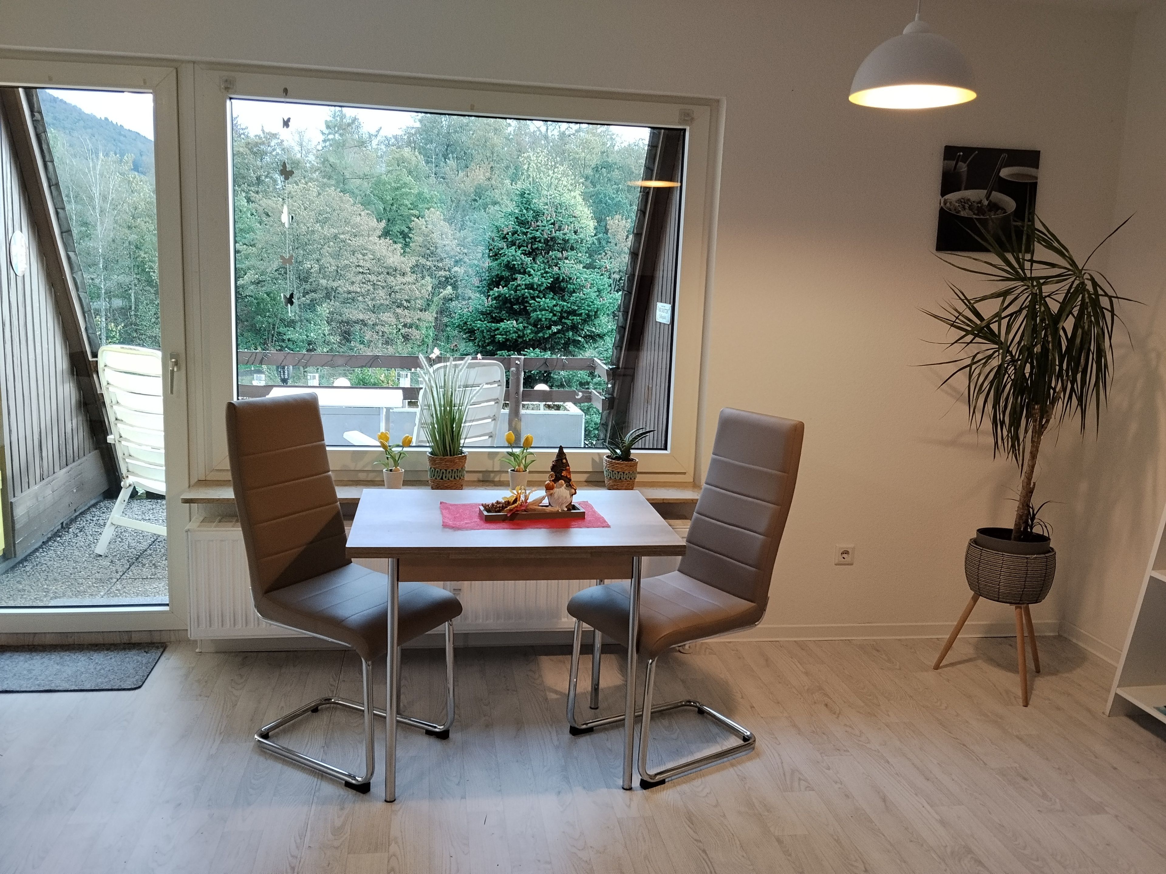 Un comedor con una mesa, sillas, una planta en maceta y vistas a un bosque a través de grandes ventanales.