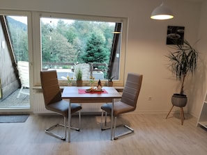 Un comedor con una mesa, sillas, una planta en maceta y vistas a un bosque a través de grandes ventanales.