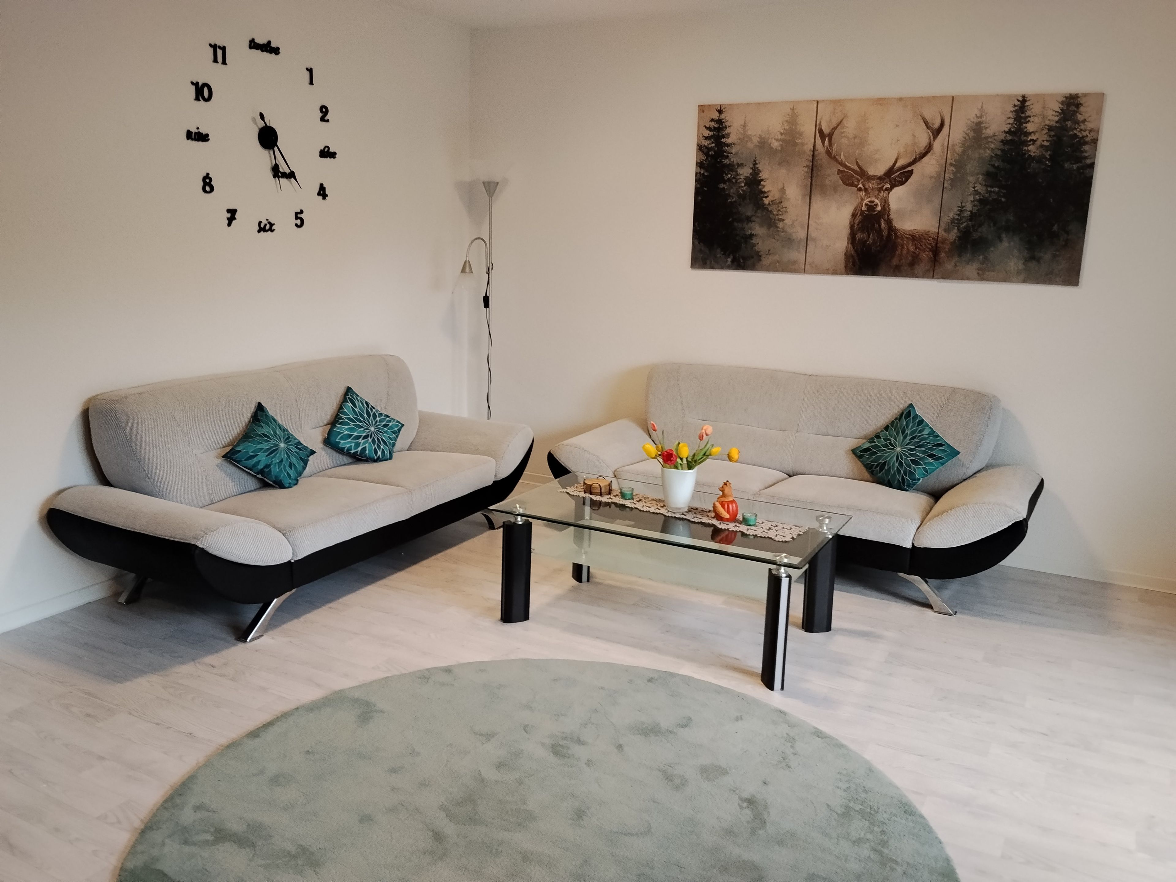 Ein Wohnzimmer mit zwei Sofas, einem Couchtisch, einer Uhr und einem Wandbild zum Thema Wald.