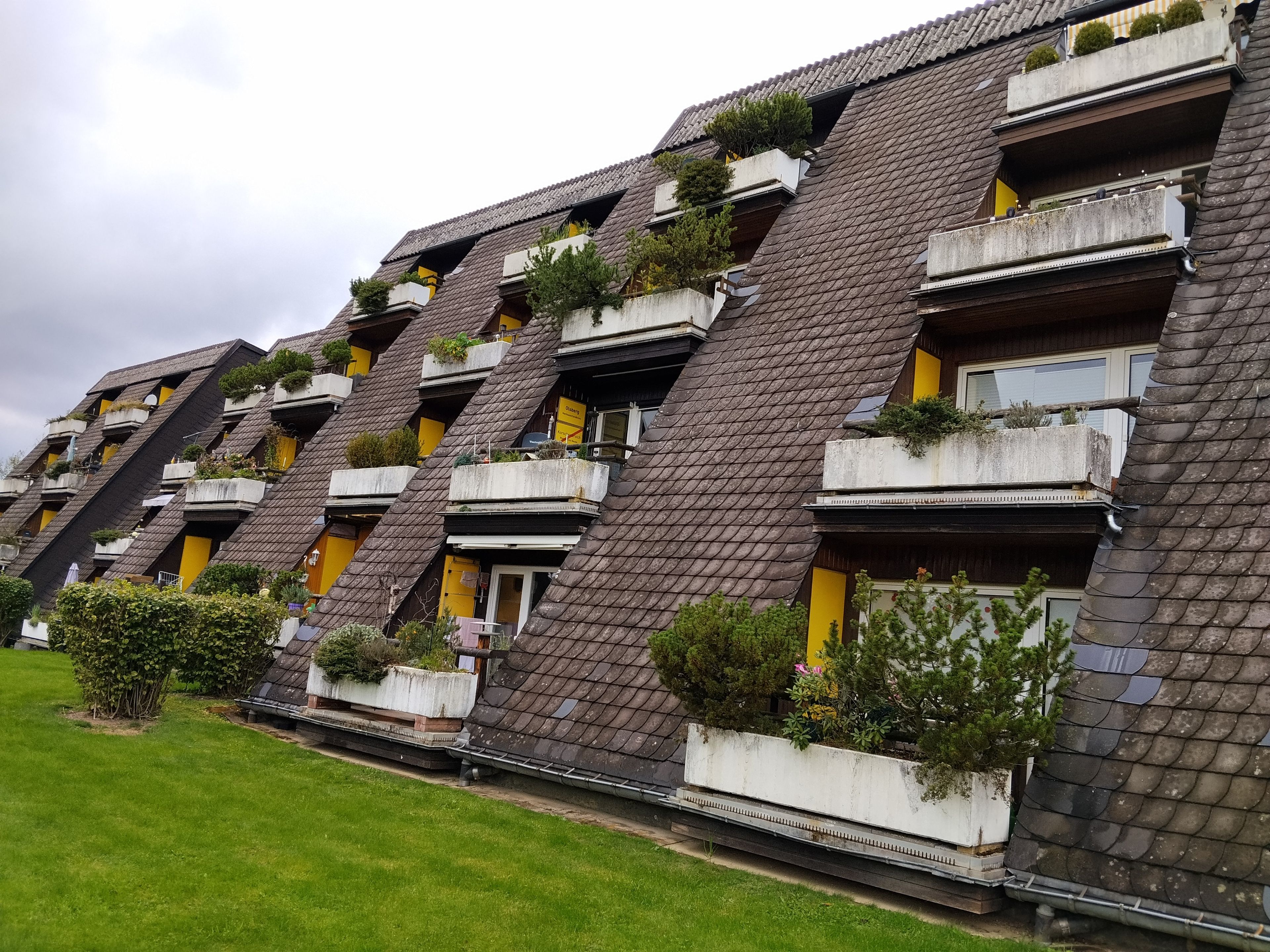Una hilera de edificios residenciales con techos inclinados, balcones con plantas y una zona verde al frente.