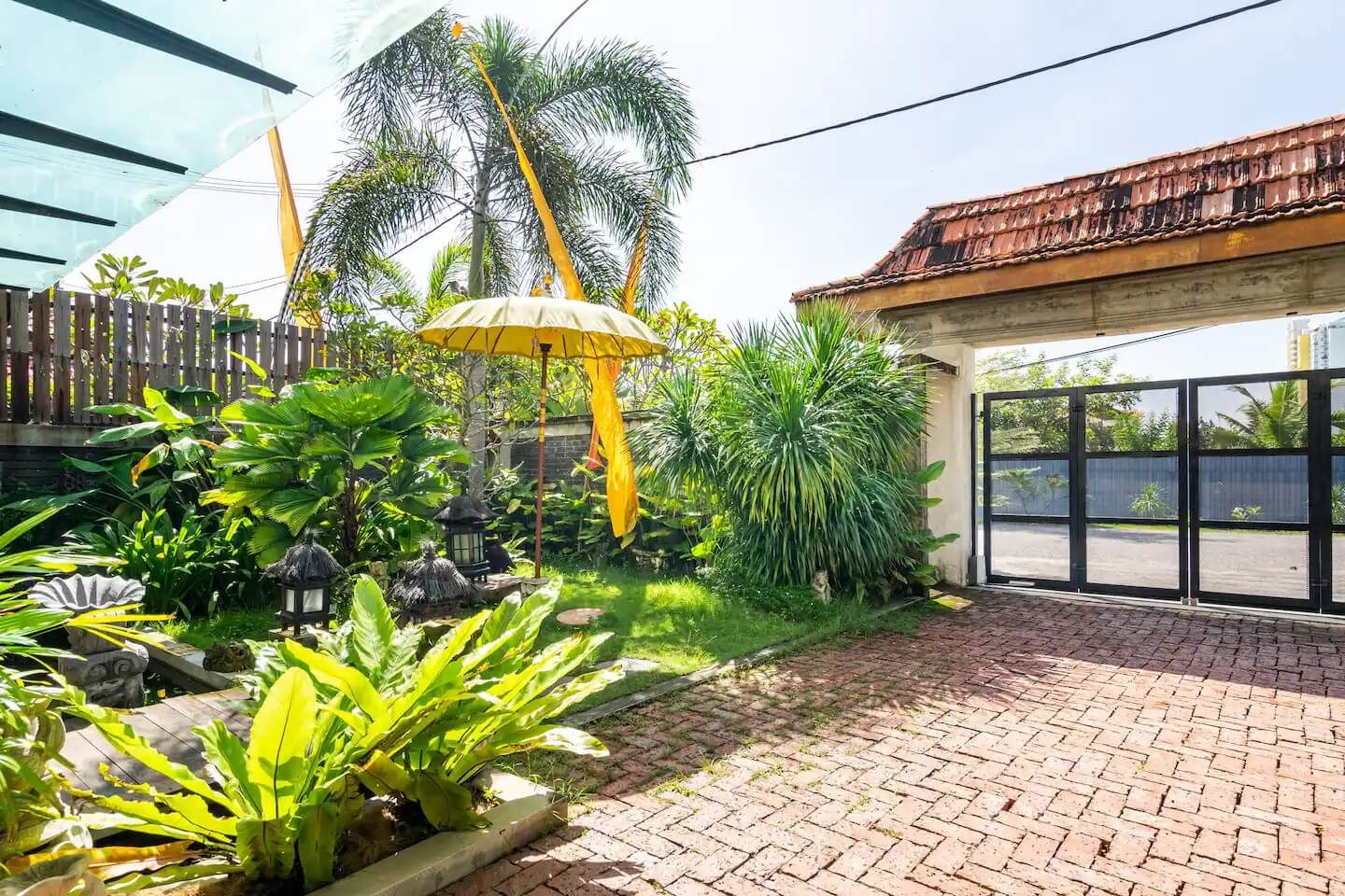 A courtyard with a brick pathway, tropical plants, a yellow umbrella, and a traditional lantern.