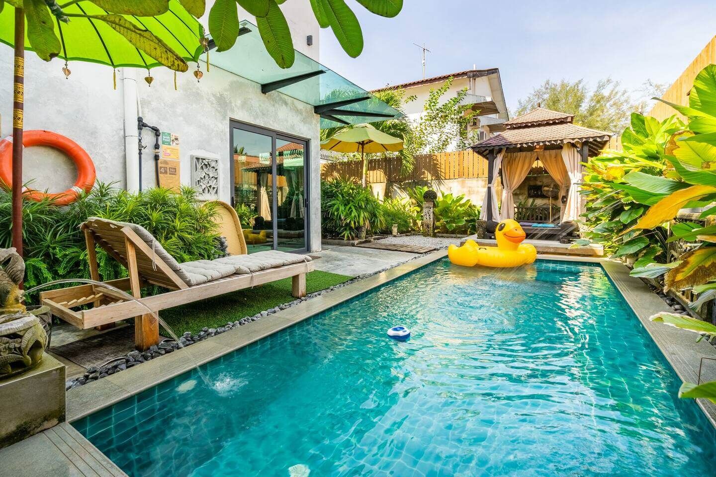 Una piscina con cubierta de madera y sillones de descanso, rodeada de exuberante follaje y una casa con puerta de cristal.