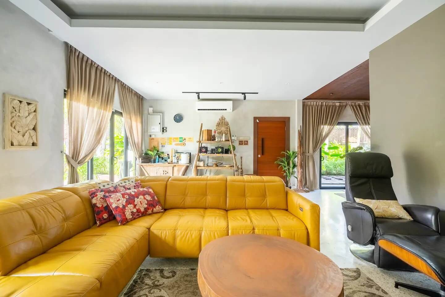 A living room with a yellow sofa, a black chair, and a coffee table. There is a kitchen area with a wooden door and a shelf with various items.