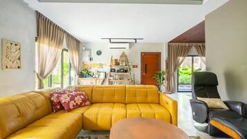 A living room with a yellow sofa, a black chair, and a coffee table. There is a kitchen area with a wooden door and a shelf with various items.